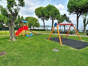 Un parque infantil con un tobogán rojo y columpios, frente a un fondo de árboles y una masa de agua.