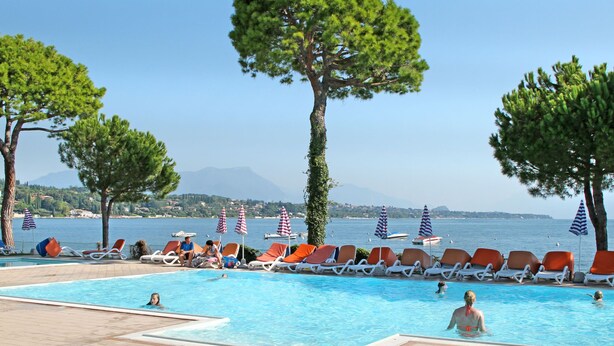 A poolside area with lounge chairs and umbrellas, a clear view of the sea, and a mountainous backdrop.