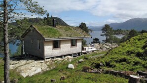 Une cabane rustique à toit de chaume, située sur une colline verdoyante, avec un lac et des montagnes en arrière-plan.
