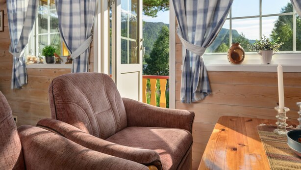 A cozy room with a wooden interior, a comfortable chair, a table with a candle, and a view of greenery through large windows. - Synnøve by Interhome (Hellevik I Fjaler)