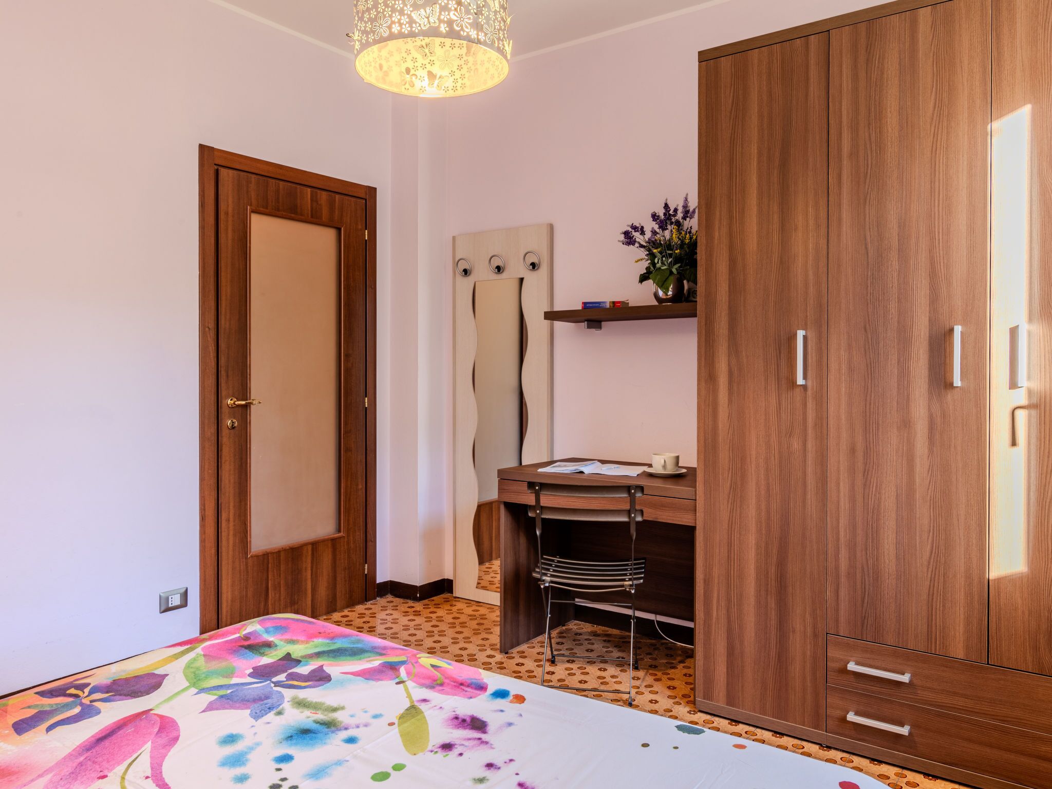A bedroom with a bed, wooden wardrobe, desk, chair, and a mirror.