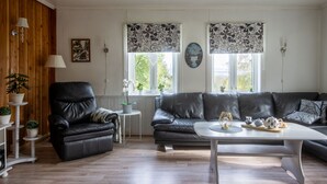A living room with a black leather sofa, a white coffee table, and a black armchair. There are two windows with patterned curtains, a white lamp, and a small table with plants. - Gulehuset by Interhome (Reksteren)