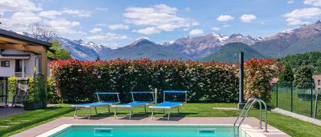 Una piscina con tres sillas reclinables azules, un seto con flores rojas y montañas al fondo.