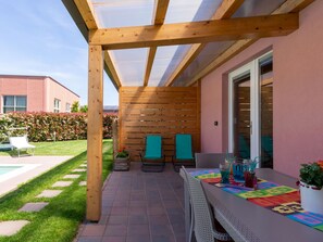 An outdoor patio with a wooden pergola, a table with colorful placemats, two chairs, and a potted plant. - Gelsomini (CCO521) by Interhome (Colico)