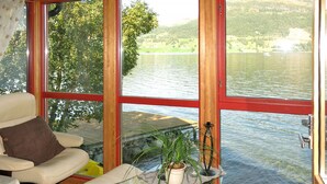 Ein Zimmer mit großem Fenster mit Seeblick, ein Sessel mit Kissen, ein Glastisch mit Pflanze und ein Fahrrad, das an das Fenster gelehnt ist.