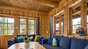 A cozy wooden cabin interior with a large wooden table, a sofa with cushions, and a window with a view of trees.