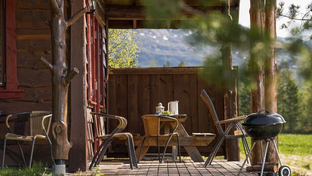 A rustic wooden cabin with a porch, wicker chairs, a table with a kettle, and a grill.