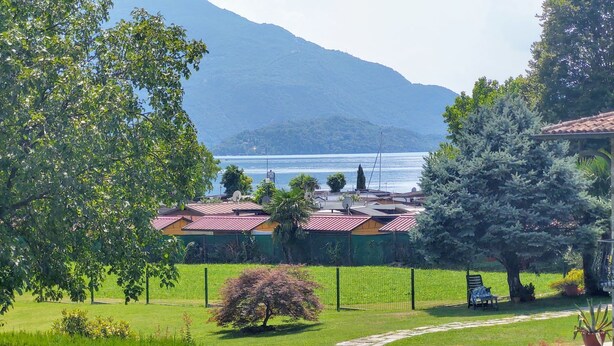 A residential area with a view of a lake and mountains. There are houses with red roofs, a stone gate, and a well-maintained lawn.