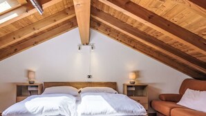 An attic bedroom with a wooden ceiling, a bed with white bedding, two bedside tables with lamps, and a brown sofa.