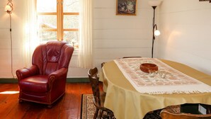 Una habitación con suelo de madera, un sillón de cuero rojo, una mesa con mantel, una silla de madera, una lámpara y una ventana con cortinas.