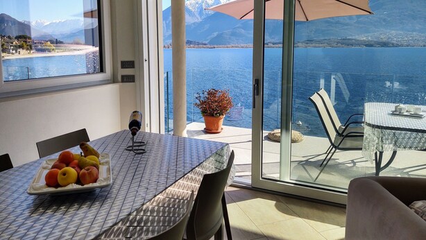 A dining area with a table set for two, a potted plant, and a view of a lake with mountains in the background. - Gelsomino (GLA125) by Interhome (Gera Lario)