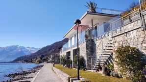 A coastal building with a stone facade, a balcony with a glass railing, and a view of the sea and mountains in the background. - Gelsomino (GLA125) by Interhome (Gera Lario)