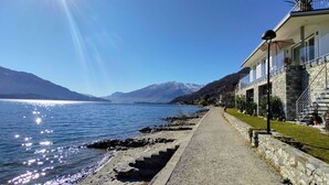 A coastal building with a stone facade, a paved walkway, and a clear view of the lake and mountains in the background. - Gelsomino (GLA125) by Interhome (Gera Lario)