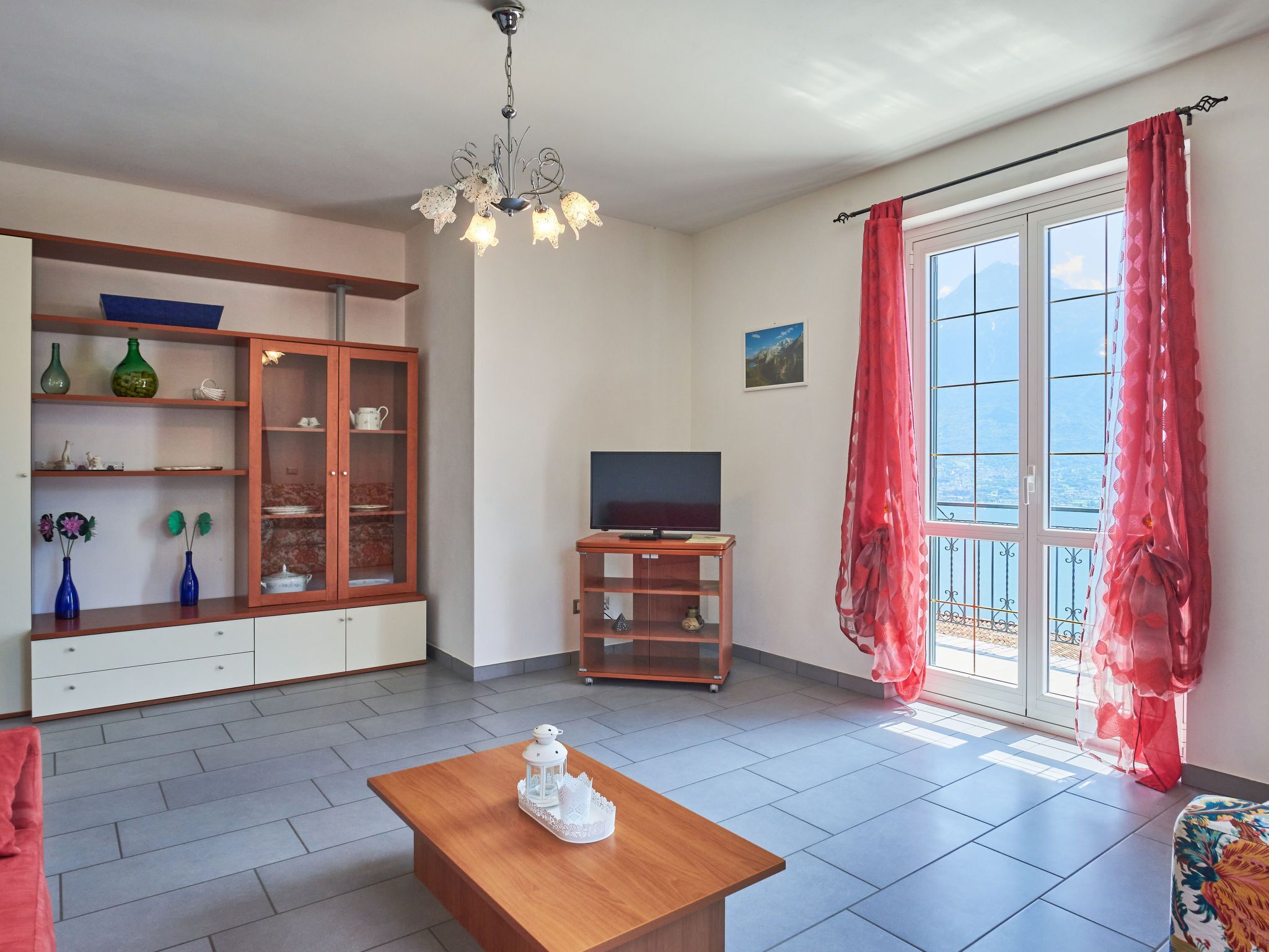 A living room with a wooden coffee table, a TV on a stand, a cabinet with glass doors, and a large window with red curtains.