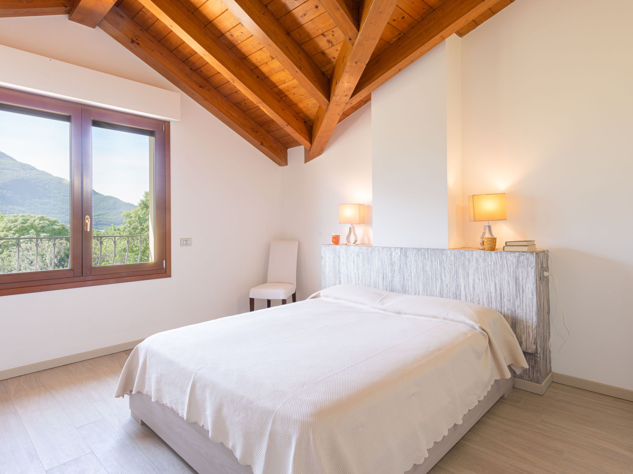A bedroom with a large bed, wooden ceiling beams, a window with a view of greenery, and two bedside lamps.