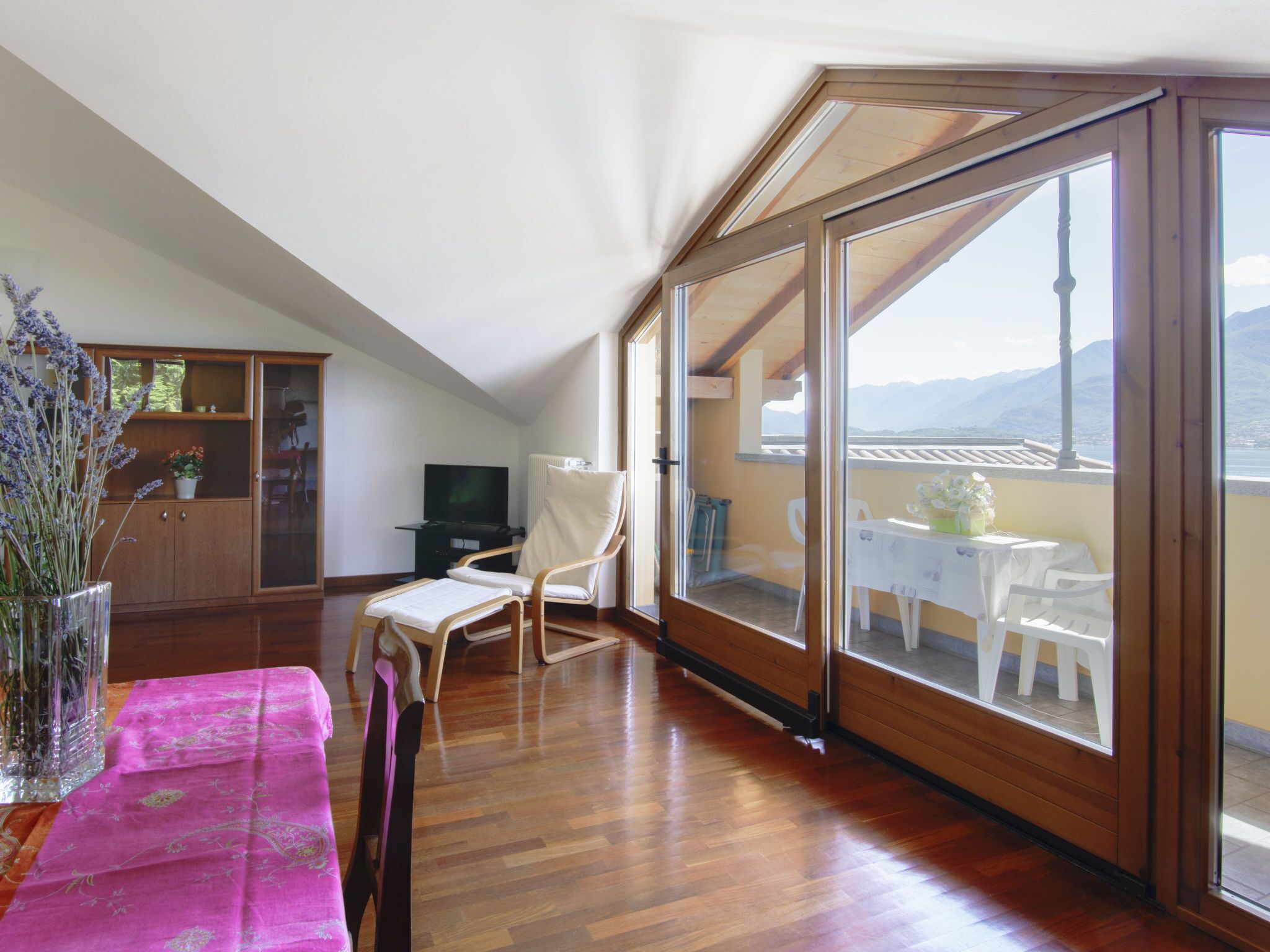 A room with a wooden floor, a dining table with chairs, a TV, a cabinet, and a large glass door leading to an outdoor balcony with a table and chairs.