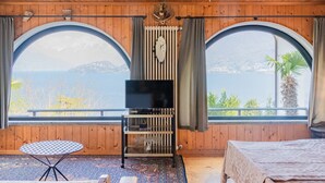 Une pièce chaleureuse avec un plafond en bois, un téléviseur posé sur un meuble, un tapis à motifs, deux lits et de grandes fenêtres en arc d’où l’on peut admirer un paysage magnifique.