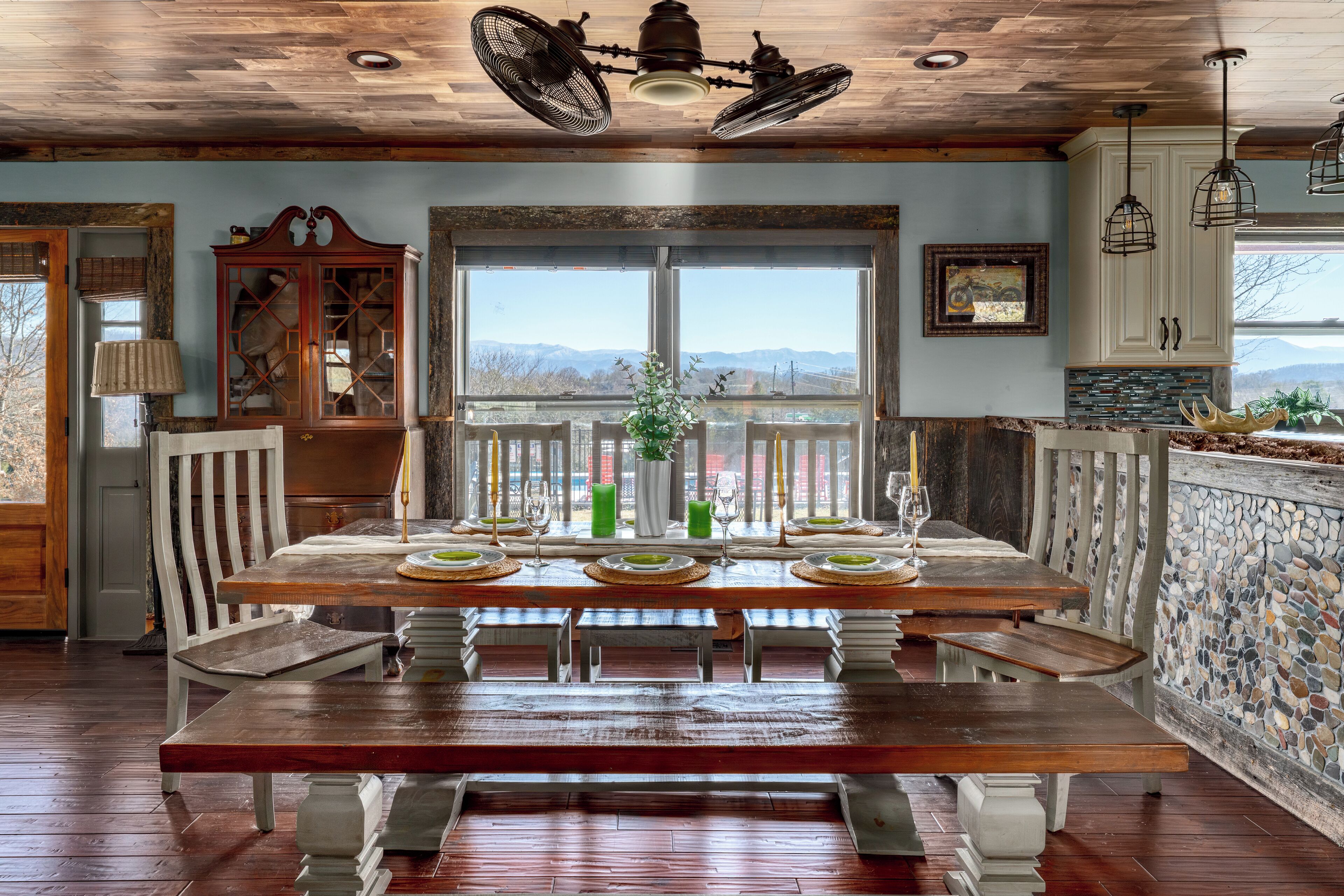 A dining area with a wooden table set for six, a large window with a view, and a stone wall.
