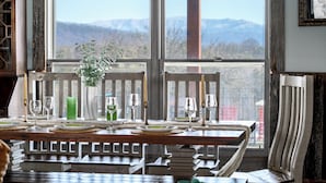 Mesa de comedor servida con platos, vasos y un florero con follaje, ubicada cerca de una ventana con vista a la montaña.