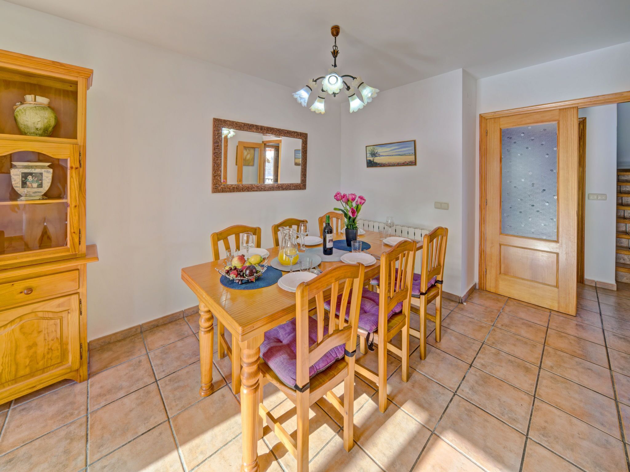 Un coin repas comprenant une table en bois, des chaises, un lustre, un miroir et un tableau accroché au mur.