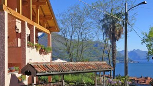 A building with a wooden balcony and a tiled roof, surrounded by greenery and a view of a lake and mountains. - Ruscello by Interhome (San Siro)