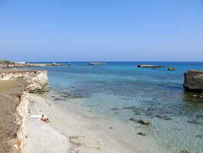 A beach with clear water, rocky outcrops, and a person lying on the sand.