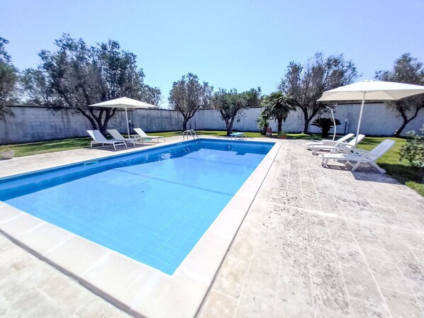A rectangular swimming pool with a tiled edge, surrounded by a tiled deck. There are lounge chairs and umbrellas along the poolside.