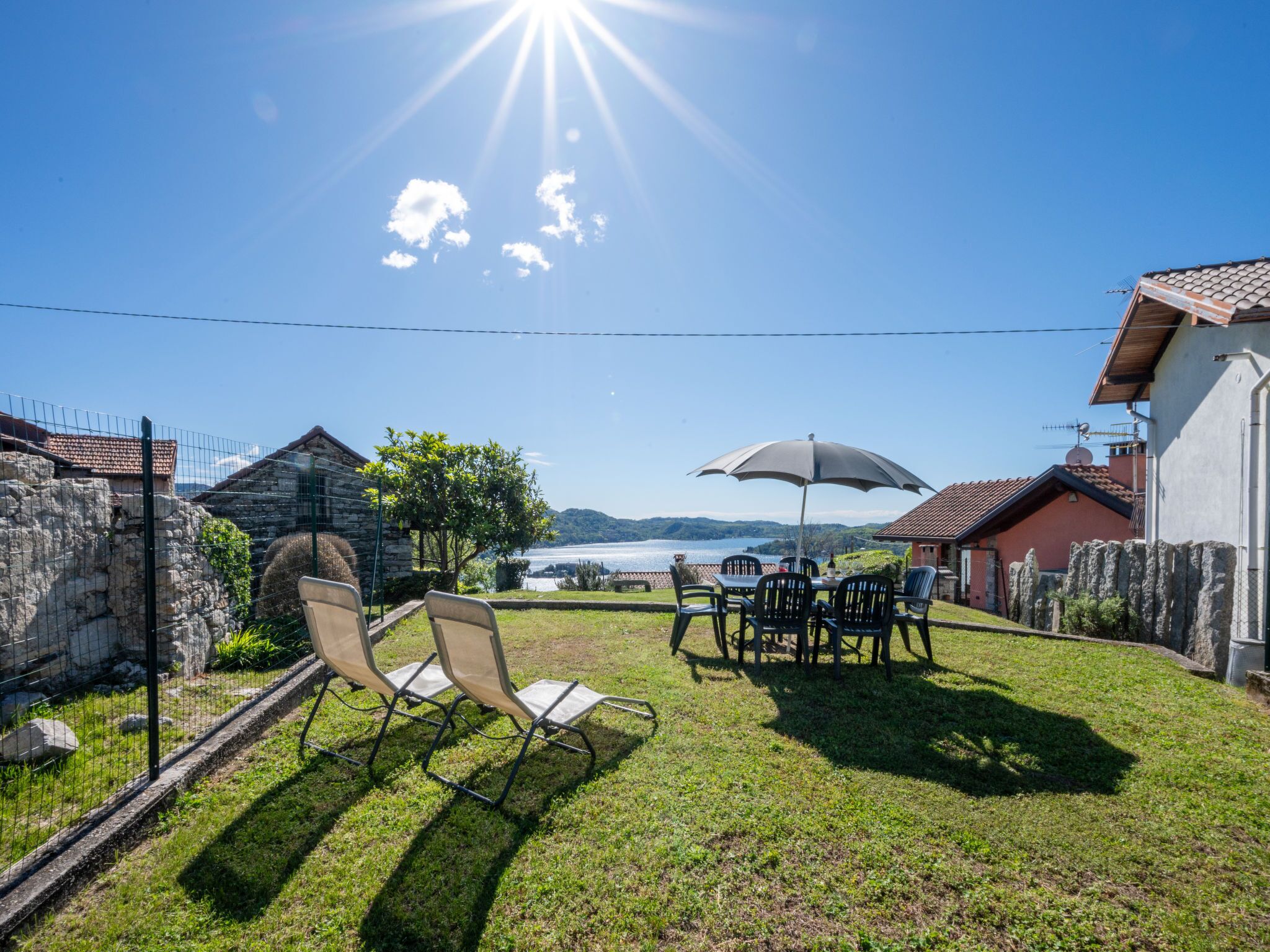 A sunny outdoor seating area with a table and chairs, a large umbrella, and a view of a lake and mountains in the background.