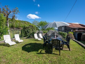 An outdoor seating area with lounge chairs and a table under a large umbrella, surrounded by greenery and a stone building. - Monte San Giulio by Interhome (Orta San Giulio)