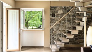 Un intérieur moderne avec un escalier en colimaçon adossé à un mur en pierre, une grande fenêtre donnant sur des arbres et un lustre.
