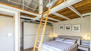 Une chambre avec une échelle en bois, un lit avec une literie à rayures, une table de chevet avec une lampe et des tableaux encadrés accrochés au mur.
