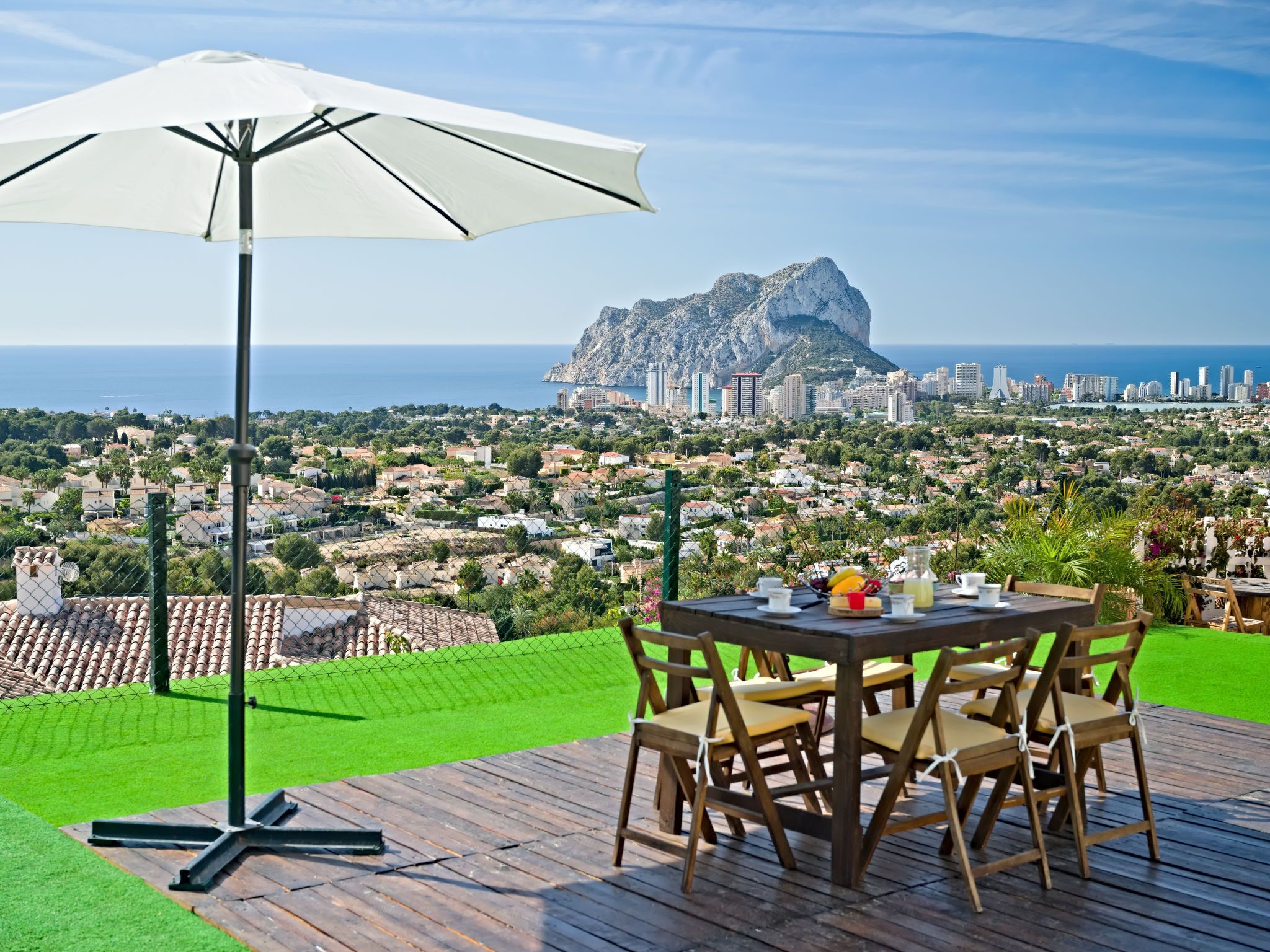 A table with chairs and a white umbrella on a wooden deck overlooking a coastal cityscape with a prominent rocky outcrop.