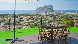 Een tafel met stoelen en een witte parasol op een houten terras met uitzicht op een kustplaats en een opvallende rotsformatie.