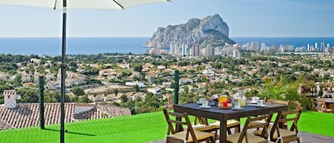 A table with chairs and a white umbrella on a wooden deck overlooking a coastal cityscape with a prominent rocky outcrop.