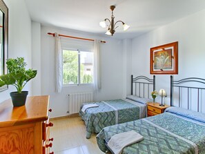 A bedroom with two beds, a radiator, a window with curtains, a potted plant, a wooden dresser, a nightstand with a lamp, and a framed picture on the wall. - Altamar by Interhome (Calpe/Calp)