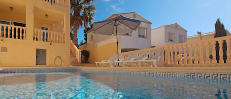 A clear swimming pool with lounge chairs and umbrellas, surrounded by buildings with balconies and palm trees.