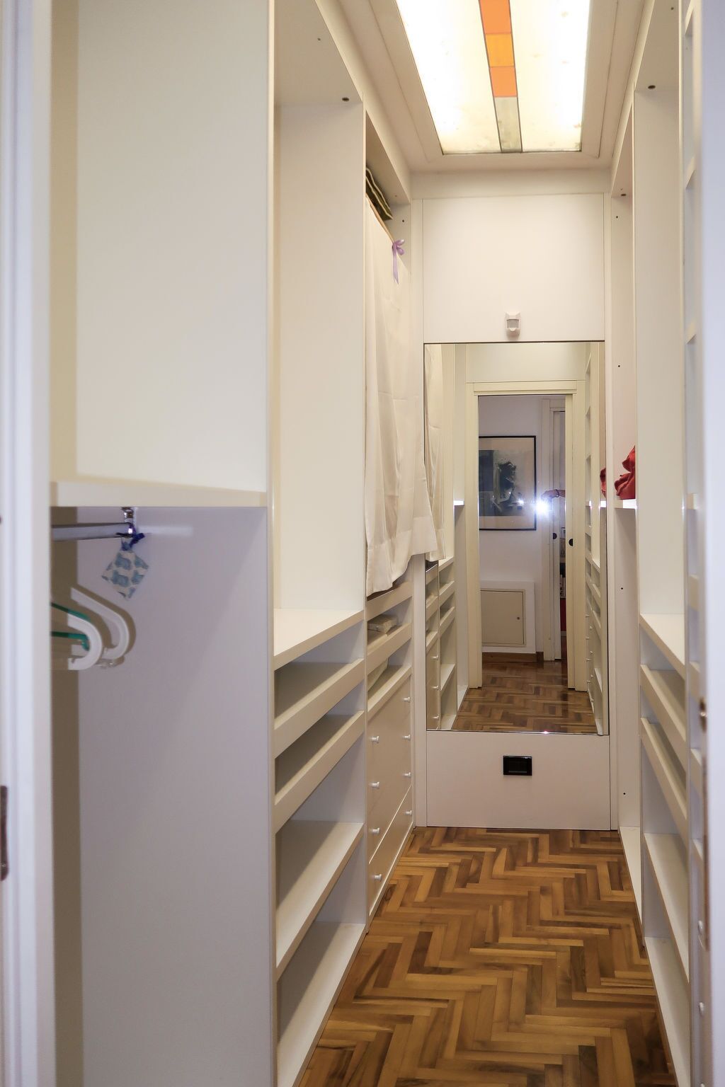Un estrecho pasillo con suelo de madera en espiga, paredes blancas y un espejo que refleja la habitación.