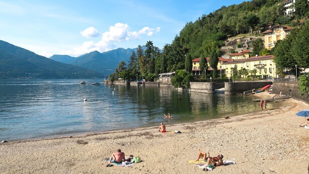 A beach with people sunbathing, a lake, and buildings in the background.