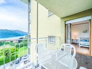 A balcony with a table and chairs, a bed with a blue and white patterned bedspread, and a view of a lake and mountains.