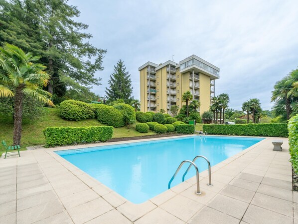A swimming pool with a clear blue water, surrounded by a tiled deck. In the background, there is a multi-story building with balconies.