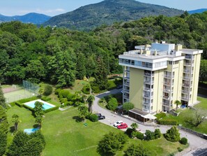 A multi-story building with balconies, surrounded by lush greenery and a tennis court.