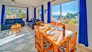 Une pièce lumineuse avec une table à manger dressée pour deux personnes, un canapé et une vue sur la mer.