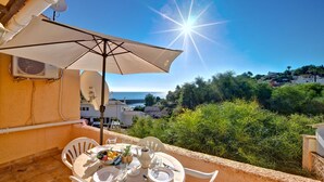Une terrasse avec une table dressée pour deux personnes, un parasol blanc et une vue sur la mer et la verdure.