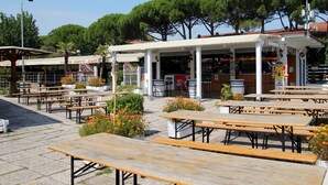An outdoor café with wooden tables and benches, surrounded by trees and a building with a tiled roof.