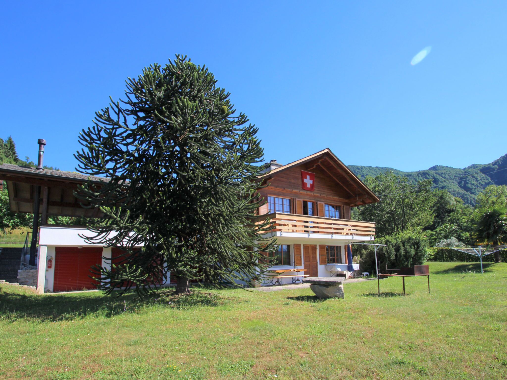 A two-story wooden chalet with a Swiss flag, a large tree in front, and a mountain backdrop.