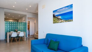 Ein Wohnzimmer mit blauem Sofa, einem Essbereich mit Tisch und Stühlen sowie einem an der Wand hängenden Bild von einer Küstenlandschaft.
