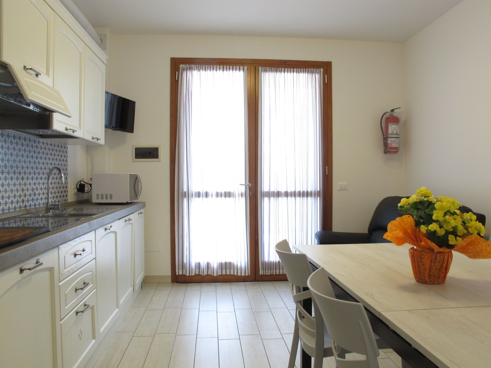 Una cocina moderna con gabinetes blancos, un microondas y un lavamanos. Hay una zona de comedor con una mesa y sillas, y un extintor de incendios montado en la pared.
