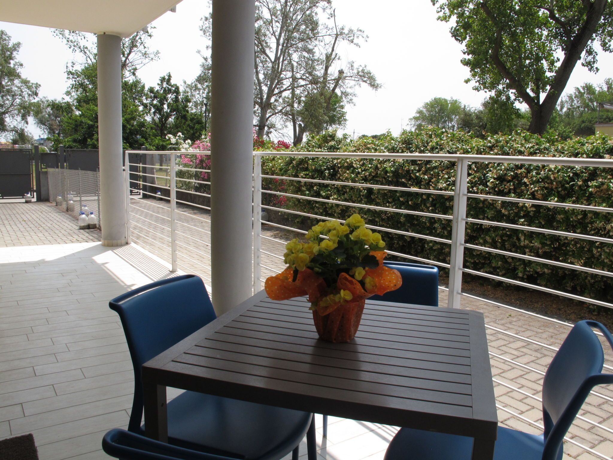 A covered outdoor seating area with a table and chairs, a flower vase, and a view of a hedge and trees.