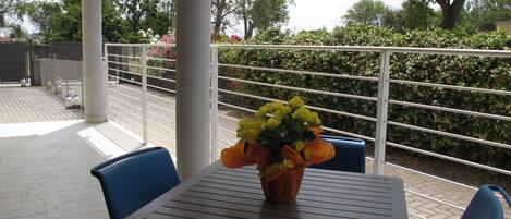 A covered outdoor seating area with a table and chairs, a flower vase, and a view of a hedge and trees.