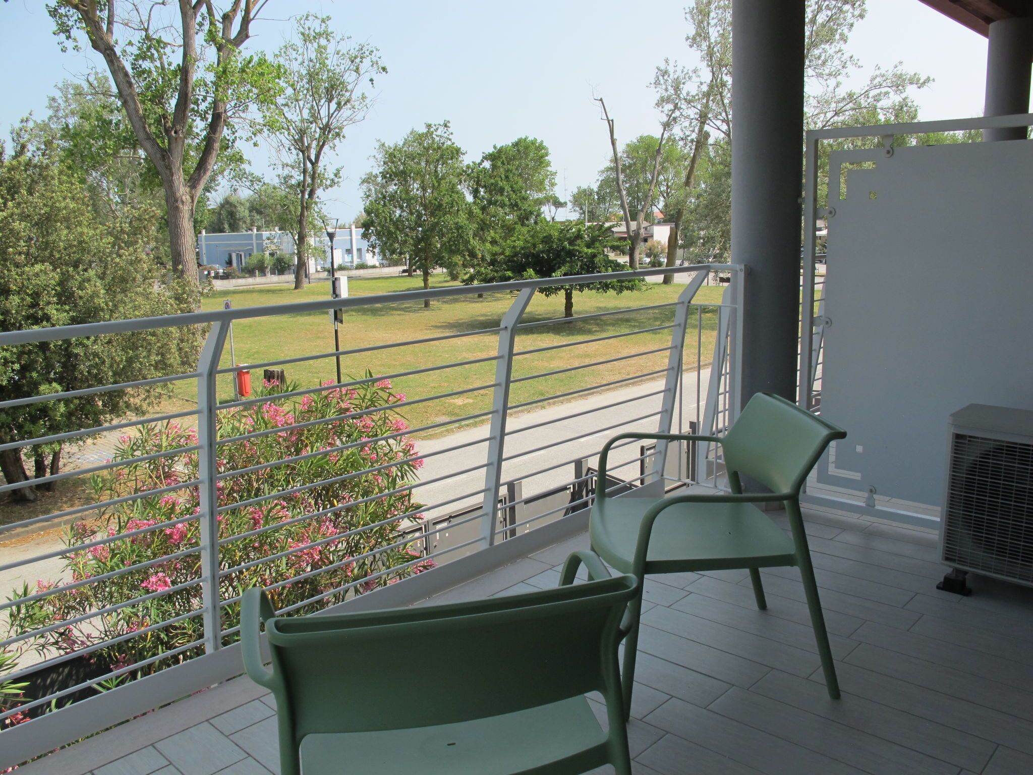 Een balkon met twee groene stoelen, een metalen hekwerk en uitzicht op een grasveld met bomen en een gebouw op de achtergrond.